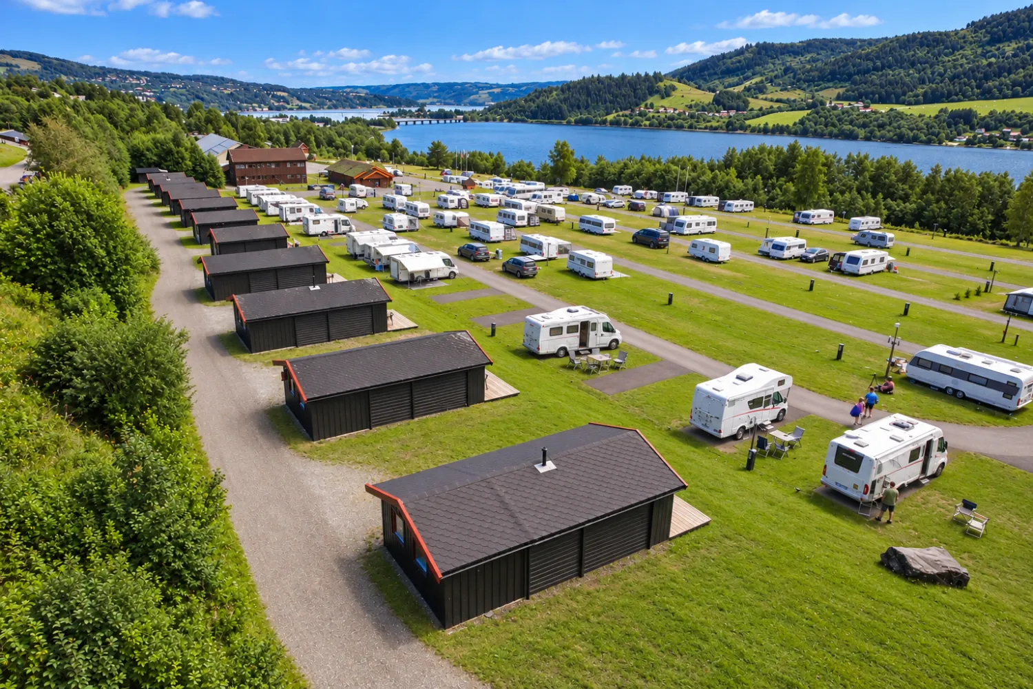 Utsikt over campingplass med hytter og bobiler på gress, ved en blå innsjø og grønne åser under blå himmel.