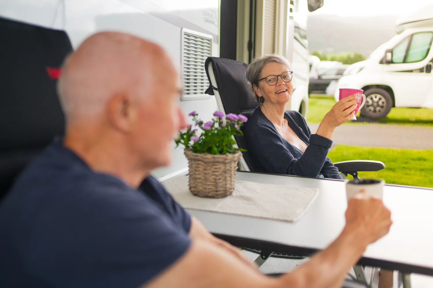 Kvinne sitter ved et bord utenfor en bobil og løfter en rosa kopp, med mann i forgrunnen.