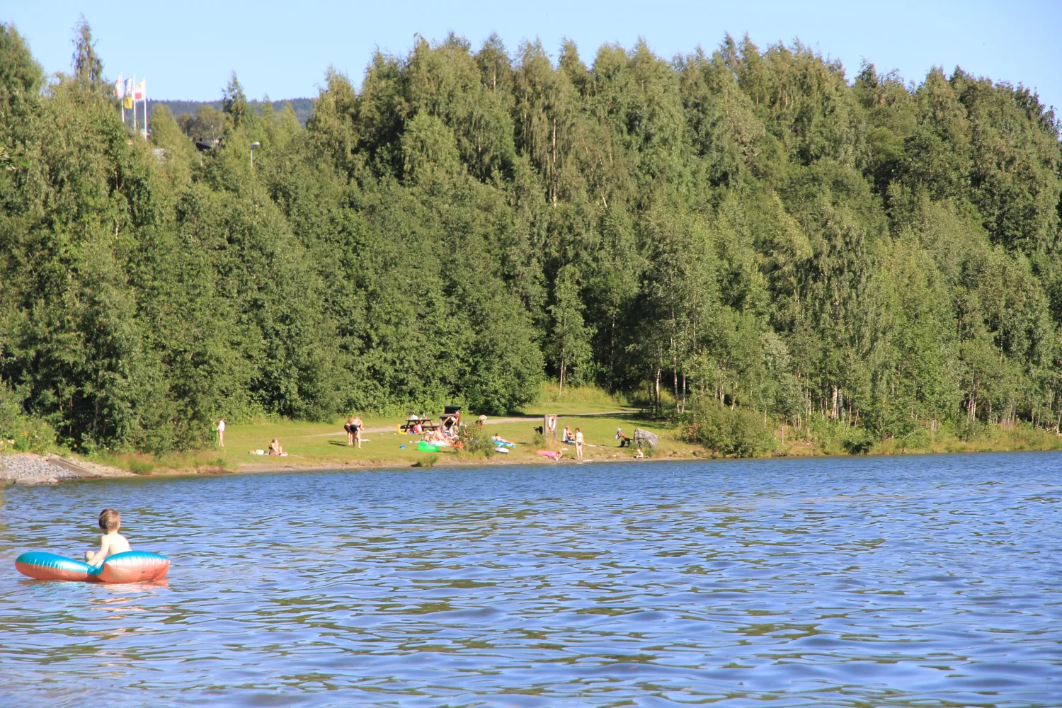 Person flyter på en bade-ring i en rolig innsjø, med grønn skog og folk på en strand og Lillehammer Turistsenter i bakgrunnen.