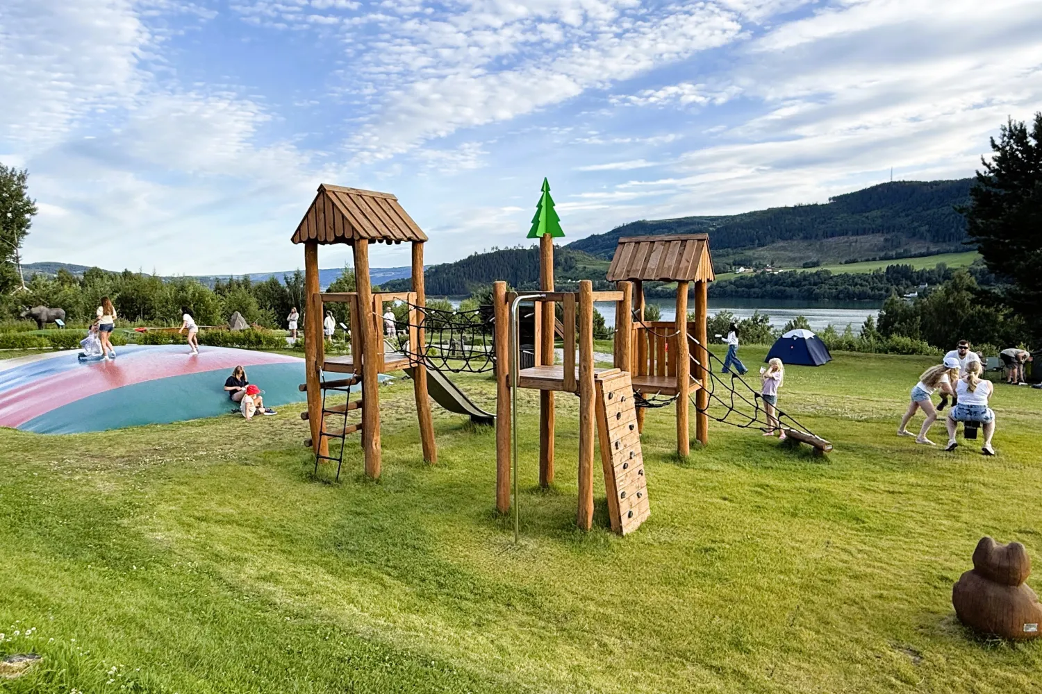 Trebasert lekeapparat på gress med barn som leker, ved en innsjø og skogkledde åser under skyet blå himmel.