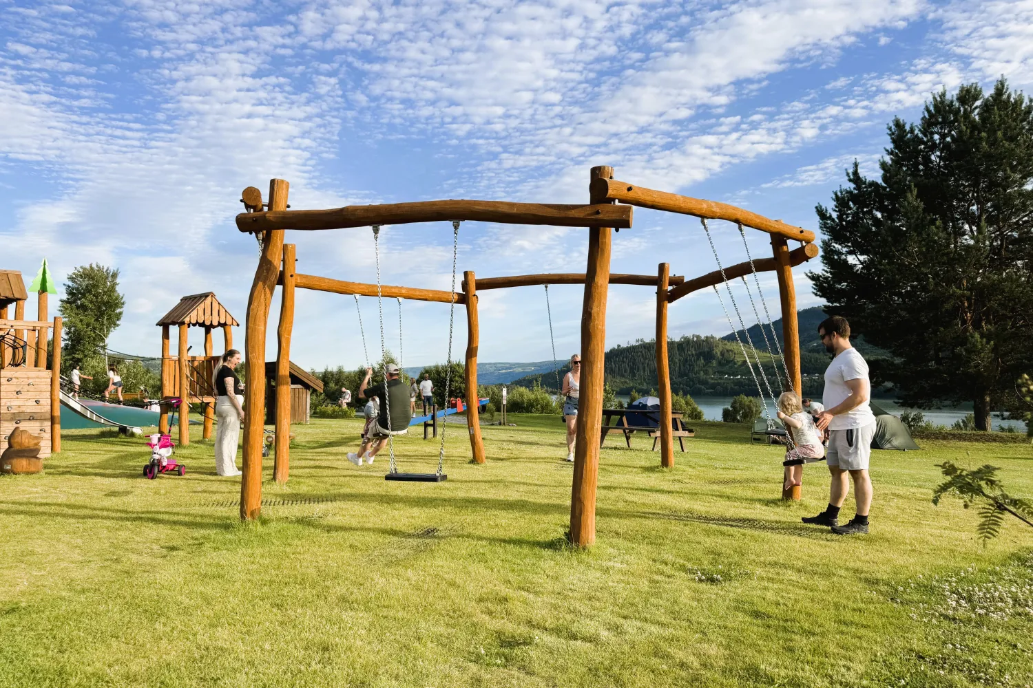 Familier på en gresskledd lekeplass med trehusker og klatrestativ, med trær, vann og skyet blå himmel.