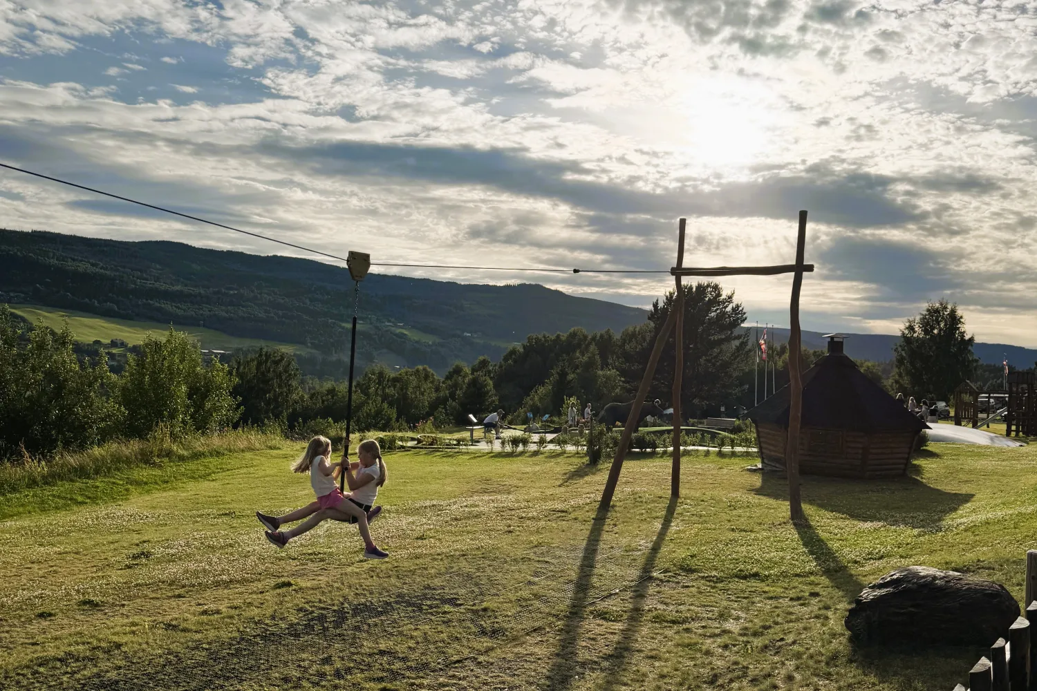 To barn suser i zipline over en gresslette, med fjell, trær og lav sol bak spredte skyer.