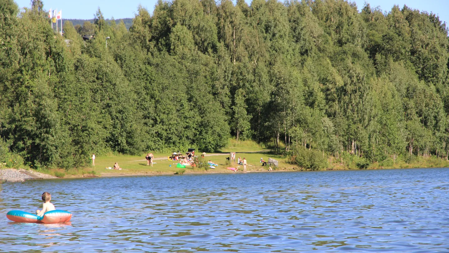 Person flyter på en bade-ring i en rolig innsjø, med grønn skog og folk på en strand og Lillehammer Turistsenter i bakgrunnen.